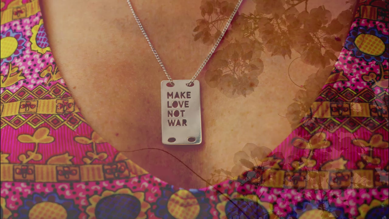 Load video: Close up on a MAKE LOVE NOT WAR dogtag pendant in silver on a chain worn by a woman